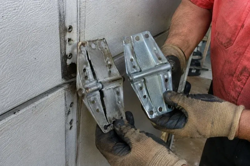 Gloved hands holding two metal garage-door hinges side by side, one discolored with a visible crack and the other bright silver. — Garage Door Hinge Replacement, Kitchener
