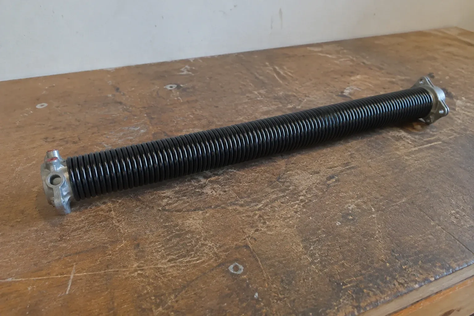 Detached black coil spring with metal end fittings on a workbench. — Torsion Spring Replacement, Kitchener